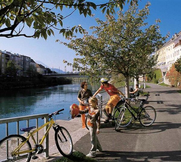 Drei Personen mit Fahrrädern und ein Kind stehen auf einem von Bäumen gesäumten Uferweg, im Hintergrund sind Gebäude und eine Brücke zu sehen.