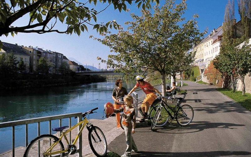 Drei Personen mit Fahrrädern und ein Kind stehen auf einem von Bäumen gesäumten Uferweg, im Hintergrund sind Gebäude und eine Brücke zu sehen.