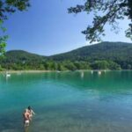Zwei Menschen stehen im flachen, klaren Wasser eines Sees, der von grünen Bäumen und bewaldeten Hügeln umgeben ist, unter einem blauen Himmel.