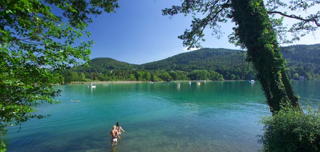 Zwei Menschen stehen im flachen, klaren Wasser eines Sees, der von grünen Bäumen und bewaldeten Hügeln umgeben ist, unter einem blauen Himmel.