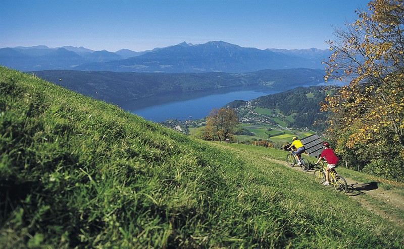 Zwei Personen fahren mit ihren Fahrrädern einen grasbewachsenen Hügel hinunter und überblicken einen See, ein Dorf und die Berge unter einem klaren blauen Himmel.