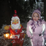 Ein kleines Kind in Winterkleidung steht neben einer Zwergenstatue, die eine brennende Laterne hält. Auf dem Boden ist Schnee zu sehen und im Hintergrund sind Bäume zu sehen.