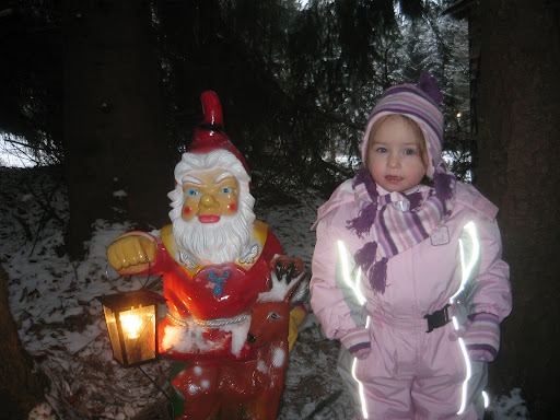 Ein kleines Kind in Winterkleidung steht neben einer Zwergenstatue, die eine brennende Laterne hält. Auf dem Boden ist Schnee zu sehen und im Hintergrund sind Bäume zu sehen.