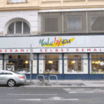Straßenansicht eines Keramikmalereistudios mit dem Schild "Keramik selbst bemalen" und geparkten Autos davor.