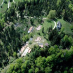 Luftaufnahme mehrerer Gebäude und einer Kapelle, umgeben von Bäumen und Grün, mit kurvenreichen Straßen, die die Landschaft durchziehen.