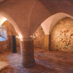 Steingewölbter Raum mit gewölbten Decken, einer zentralen Säule, einem Gitter auf dem Boden, freigelegten Ziegelwänden und gedämpfter Beleuchtung.