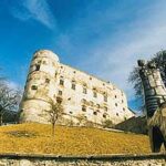 Ein steinernes Schloss auf einem grasbewachsenen Hügel ist aus einem niedrigen Winkel zu sehen, mit kahlen Bäumen und einer großen Metallskulptur im Vordergrund unter einem blauen Himmel.