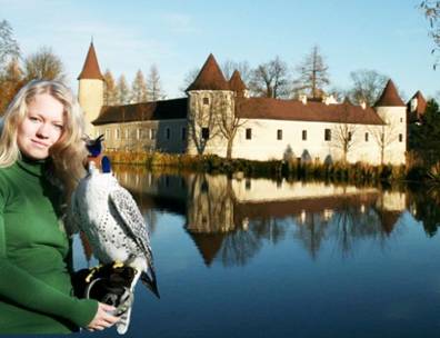 Eine Frau in einem grünen Pullover hält einen Falken an einem spiegelnden See, während im Hintergrund ein Schloss und Bäume zu sehen sind.