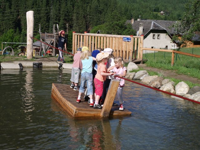 Eine Gruppe kleiner Kinder steht auf einem Holzfloß, das ein Seil hält, und überquert einen flachen Teich in einem Spielbereich im Freien mit Bäumen und Gebäuden im Hintergrund.