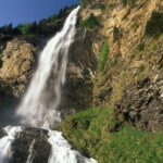 Ein hoher Wasserfall stürzt unter blauem Himmel über einen felsigen Abhang mit Vegetation und Bäumen am oberen und unteren Ende.