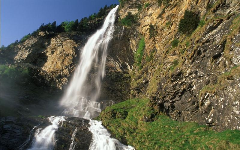 Ein hoher Wasserfall stürzt unter blauem Himmel über einen felsigen Abhang mit Vegetation und Bäumen am oberen und unteren Ende.