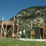 Ein hölzerner Spielplatz mit Schaukeln und Klettergerüsten liegt auf einer Wiese in der Nähe eines Wasserfalls und bewaldeter Berge unter einem klaren blauen Himmel.