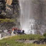 Eine Gruppe von Menschen steht auf einer steinernen Aussichtsplattform und winkt in die Kamera, während im Hintergrund ein Wasserfall und felsige Klippen zu sehen sind.