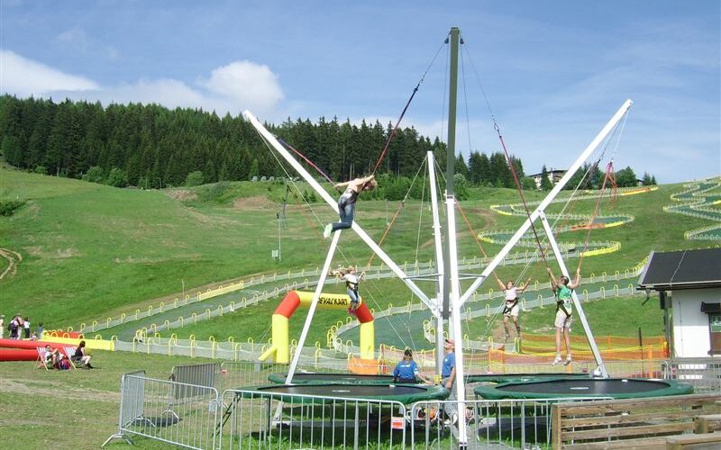 Kinder benutzen ein Bungee-Trampolin in einem Freiluftpark mit grasbewachsenen Hügeln, einem Wald und Menschen, die sich in der Nähe entspannen.