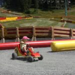 Ein Kind mit einer rosafarbenen Mütze fährt ein kleines Gokart auf einer Schotterpiste mit kurvenreichen Hindernissen und grasbewachsenen Hügeln im Hintergrund.