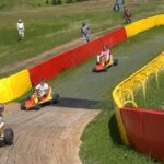 Vier Personen fahren Gokarts auf einer eingezäunten, mit roten und gelben Absperrungen versehenen Strecke, während einige Zuschauer auf einer Wiese in der Nähe zusehen.
