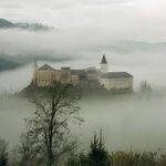 Ein großes Schloss liegt auf einem Hügel, umgeben von dichtem Nebel und Bäumen, und im Hintergrund sind bewaldete Berge zu sehen.