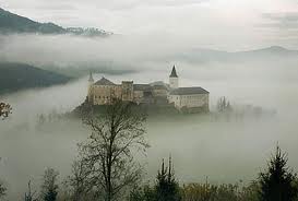 Ein großes Schloss liegt auf einem Hügel, umgeben von dichtem Nebel und Bäumen, und im Hintergrund sind bewaldete Berge zu sehen.