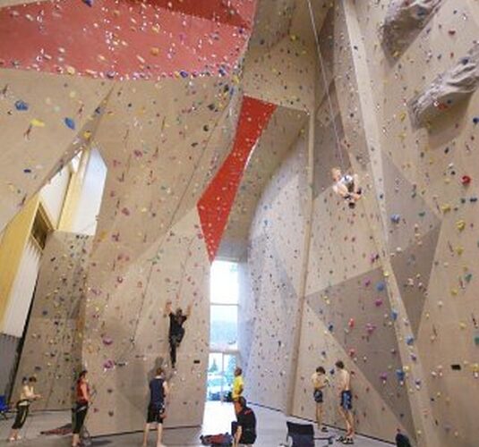Indoor-Kletterhalle mit hohen Wänden, bunten Griffen, mehreren Kletterern und anderen, die beobachten oder am Boden warten.