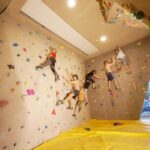 Vier Personen klettern in der Halle an einer Boulderwand mit bunten Griffen; eine gelbe Matte bedeckt den Boden zur Sicherheit.