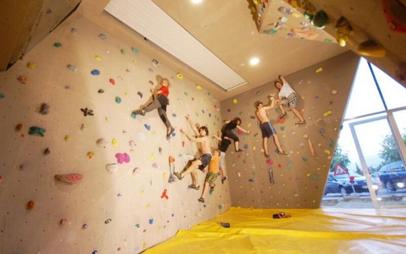 Vier Personen klettern in der Halle an einer Boulderwand mit bunten Griffen; eine gelbe Matte bedeckt den Boden zur Sicherheit.