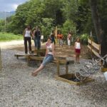 Eine Gruppe von Kindern und Erwachsenen steht und sitzt auf hölzernen Spielgeräten im Freien, umgeben von Bäumen und Kies.