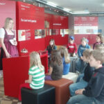 Eine Frau in traditioneller Kleidung spricht zu einer Gruppe von Kindern, die in einem Museum oder einer Ausstellung vor roten Informationsschildern sitzen.