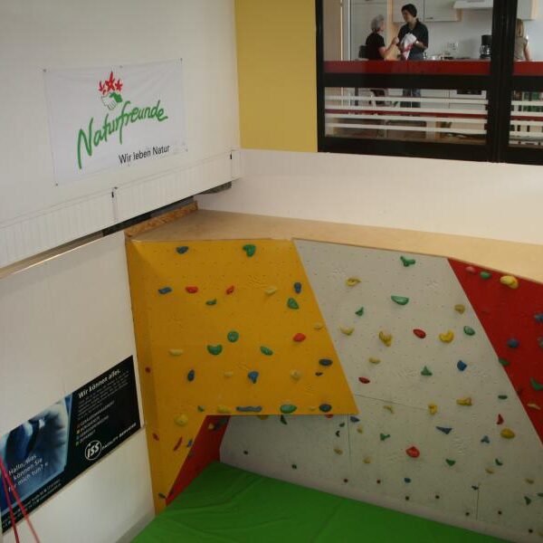 Indoor-Kletterwand mit bunten Griffen und einer grünen Matte darunter, die sich in einem Raum mit einem Naturfreunde-Banner und zwei Personen befindet, die hinter einer Glaswand im oberen Stockwerk zu sehen sind.