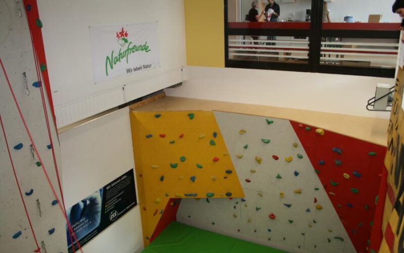 Indoor-Kletterwand mit bunten Griffen und einer grünen Matte darunter, die sich in einem Raum mit einem Naturfreunde-Banner und zwei Personen befindet, die hinter einer Glaswand im oberen Stockwerk zu sehen sind.