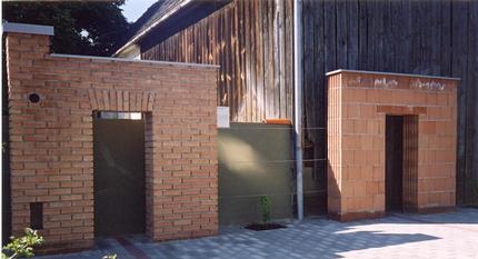 Zwei kleine Ziegel- und Schlackensteinbauten mit offenen Türen stehen neben einem Holzgebäude auf einer gepflasterten Fläche.