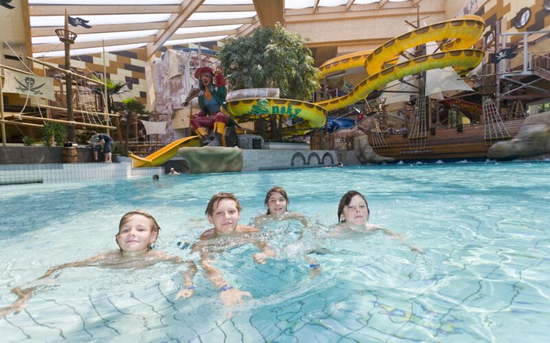Vier Kinder schwimmen in einem Hallenbad mit einer Wasserrutsche im Piratenstil und einem Spielgerät im Hintergrund.