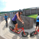 Drei Personen mit Helmen fahren auf großen Rollern auf einer hölzernen, geschwungenen Bahn im Freien, umgeben von Bäumen und Grün.