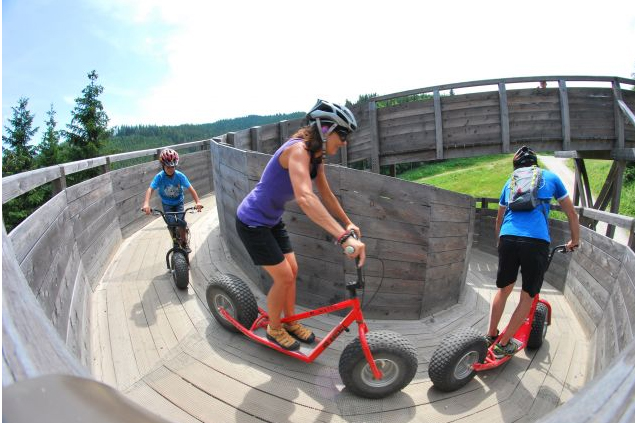Drei Personen mit Helmen fahren auf großen Rollern auf einer hölzernen, geschwungenen Bahn im Freien, umgeben von Bäumen und Grün.