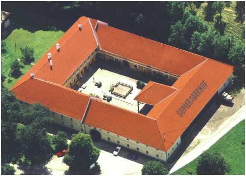 Luftaufnahme eines großen, rechteckigen Gebäudes mit rotem Ziegeldach, grünen Bäumen, die das Gebäude umgeben, und einem offenen zentralen Innenhof.