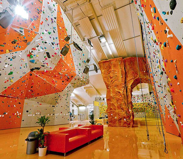 Indoor-Kletterhalle mit bunten Kletterwänden, einer künstlichen Felsstruktur, Seilen und einer roten Couch auf einem glänzenden Boden.