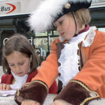 Zwei Kinder in historischen Kostümen stehen vor einem StadtBus; ein Kind liest, während das andere zuschaut, beide tragen schmucke Jacken und Rüschenkragen.
