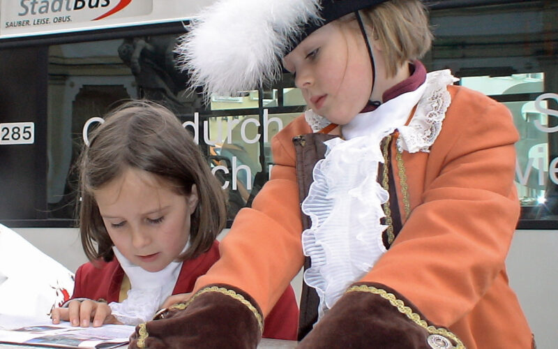 Zwei Kinder in historischen Kostümen stehen vor einem StadtBus; ein Kind liest, während das andere zuschaut, beide tragen schmucke Jacken und Rüschenkragen.