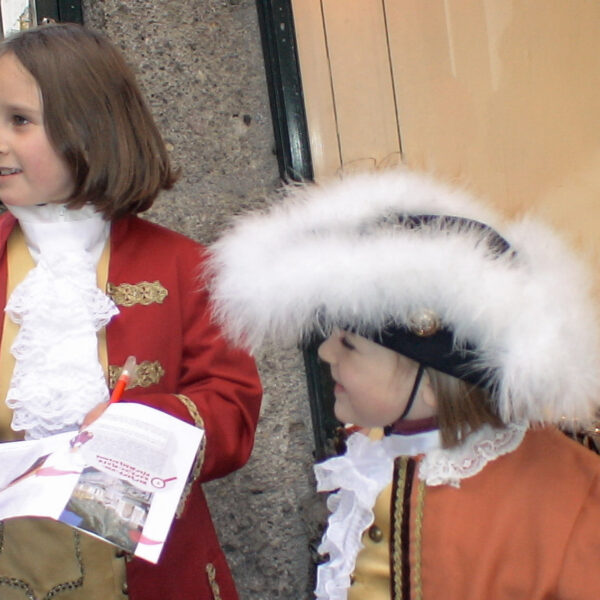 Zwei Kinder in historischen Kostümen stehen auf einem Bürgersteig; eines hält eine Broschüre in der Hand, während das andere einen großen Hut mit weißer Zierleiste trägt. Im Hintergrund ist die Auslage eines Schuhgeschäfts zu sehen.
