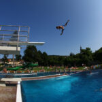 Eine Person befindet sich in der Luft, nachdem sie von einem Sprungbrett in ein Freibad gesprungen ist, während andere auf dem Sprungbrett warten und sich an einem sonnigen Tag um das Becken herum aufhalten.