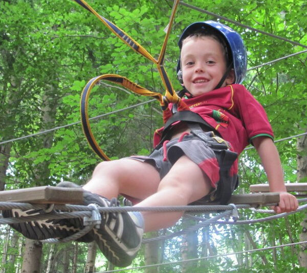 Ein Junge mit Helm und Klettergurt lächelt, während er an einem von Bäumen umgebenen Hochseilgarten teilnimmt.