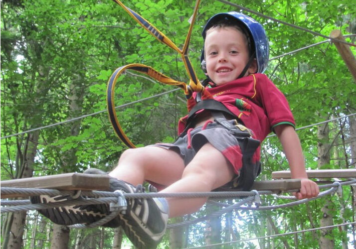 Ein Junge mit Helm und Klettergurt lächelt, während er an einem von Bäumen umgebenen Hochseilgarten teilnimmt.