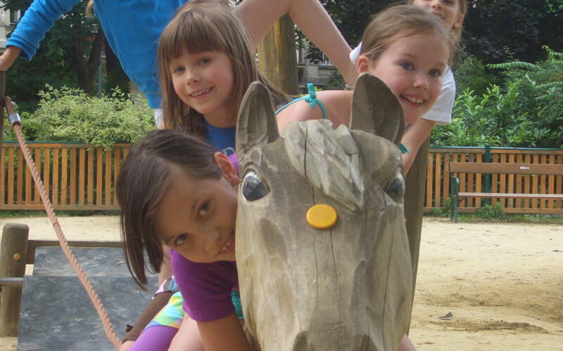 Fünf Kinder sitzen und klettern auf einem großen Holzpferd auf einem Spielplatz im Freien, lächeln und posieren für die Kamera.