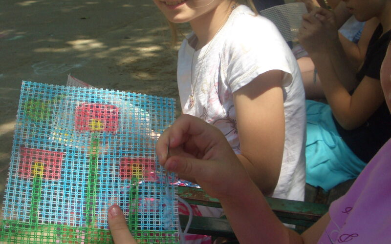 Ein Mädchen sitzt im Freien und lächelt in die Kamera, während eine andere Person ein Stück Plastikleinwand mit einem farbenfrohen Stickereimotiv in der Hand hält.