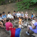 Eine Gruppe von Kindern und ein Erwachsener sitzen im Kreis auf Holzstühlen um ein Lagerfeuer in einem Waldgebiet.