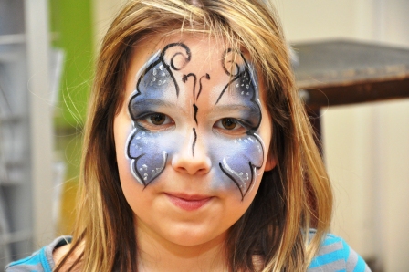 Ein junges Mädchen mit langen Haaren hat ihr Gesicht wie einen blau-schwarzen Schmetterling gemalt, mit weißen Akzenten auf Wangen und Stirn.