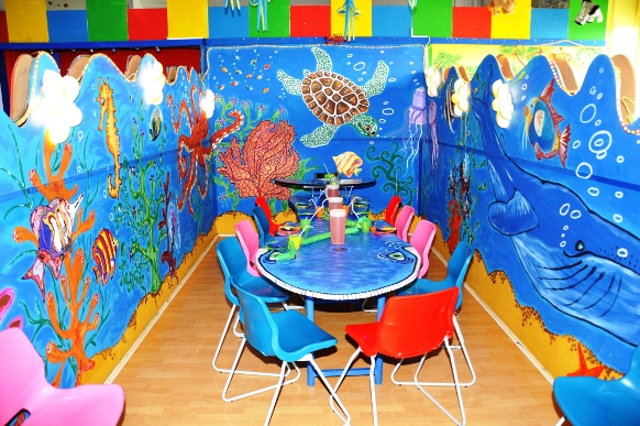 Farbenfroher Kinderpartyraum mit Unterwasserthema, mit Meeresmalereien an den Wänden, einem runden Tisch, der für eine Party gedeckt ist, und leuchtend roten, blauen und rosa Stühlen.