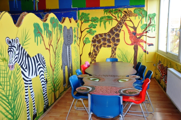Kinderspielzimmer mit einem Wandgemälde zum Thema Dschungel, das ein Zebra, einen Elefanten, eine Giraffe und einen Affen zeigt, und einem Tisch, der von bunten Stühlen umgeben ist.