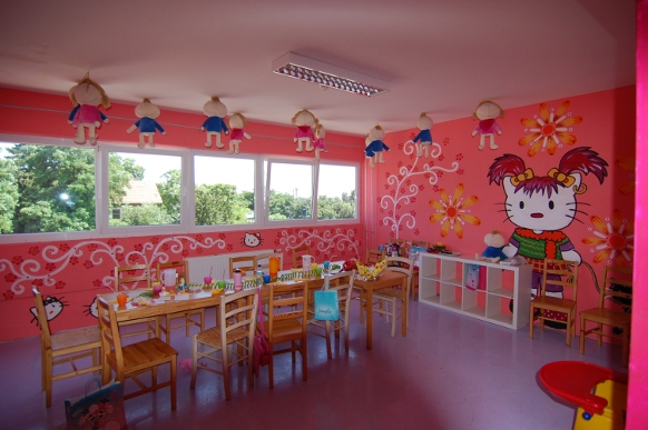 Ein farbenfrohes Klassenzimmer mit rosafarbenen Wänden, dekoriert mit Zeichentrickfiguren und Blumen, kleinen Holztischen und -stühlen und von der Decke hängenden Puppen.