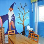 Ein Klassenzimmer mit einem Wandgemälde von einem Zauberer und einem Baum an einer blauen Wand, Holztischen und -stühlen, Truhen, einem Hochstuhl und einem Fenster, das Tageslicht hereinlässt.
