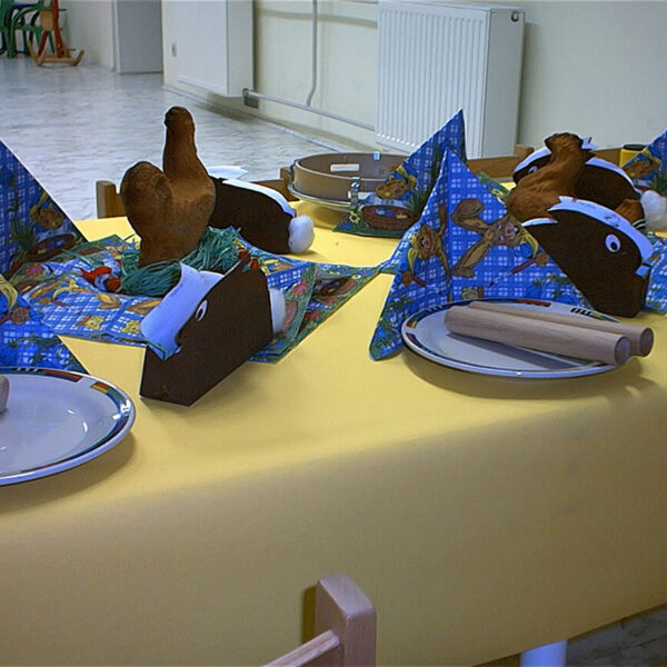 Ein gedeckter Tisch mit gelber Tischdecke, Tellern, Holzbesteck, gefalteten Servietten und Hähnchenkeulen aus Plüsch. Im Hintergrund stehen grüne Stühle und ein weißer Heizkörper.